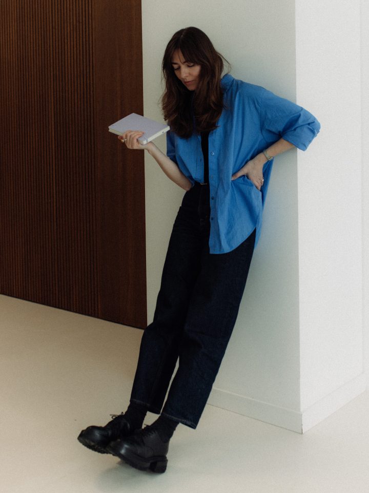 Ilona Dzen leaning against a white wall, holding a notebook
